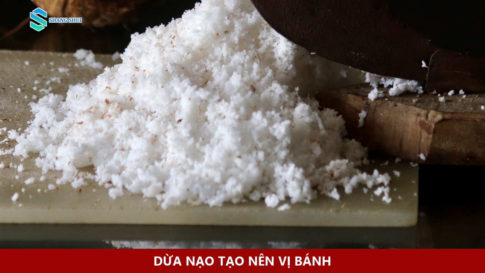 Cơm dừa nạo trắng trên thớt gỗ, cạnh phần lưỡi dụng cụ nạo.