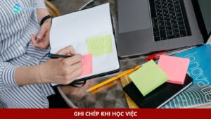 Một người viết lên giấy ghi chú đặt trong sổ bên cạnh laptop.